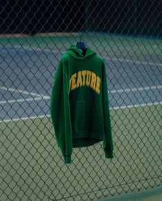 a green hoodie hangs on a chain link fence near a tennis court with the word creature printed on it