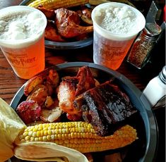 two plates with ribs, corn, and potatoes on them sitting next to beer glasses