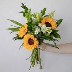 a bouquet of sunflowers and greenery in someone's hand against a white wall