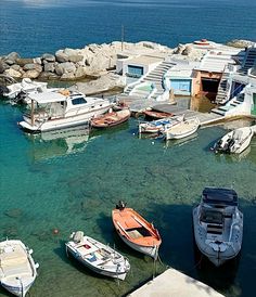 several small boats are docked in the water near some buildings and rocks, while another boat is moored to it's dock