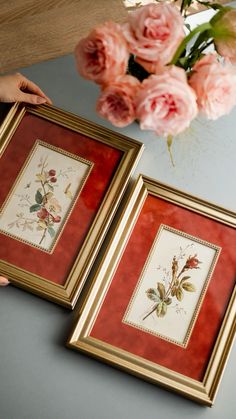 two framed pictures sitting on top of a table next to pink flowers in vases