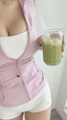 a woman in white shorts and pink jacket holding a green drink with her right hand