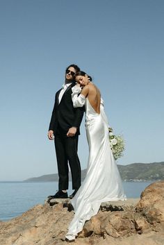 a bride and groom standing on the rocks by the water in their wedding attire, posing for a photo