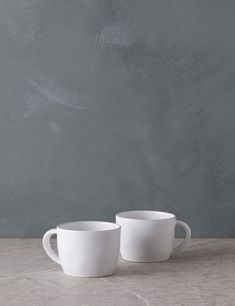 two white mugs sitting on top of a table next to each other in front of a gray wall