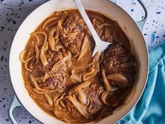 a pot filled with meat and noodles on top of a blue cloth next to a spoon