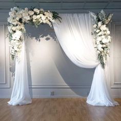 a white wedding arch decorated with flowers and greenery on a wooden floor in front of a wall