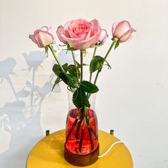 three pink roses are in a vase on a yellow table with a white wall behind it