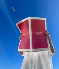 a person holding up a large piece of paper in front of their face with an airplane flying overhead