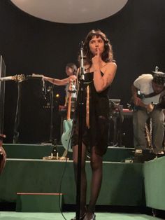 a woman standing in front of a microphone on top of a green stage with other people behind her