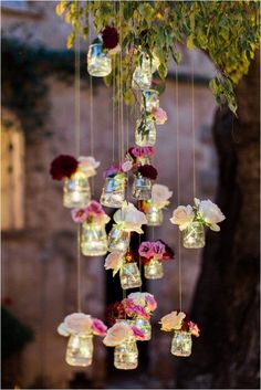 an arrangement of mason jars filled with flowers hanging from a tree in front of a building