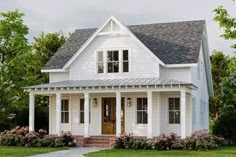 a white house with a porch and two windows on the first floor is surrounded by bushes and flowers