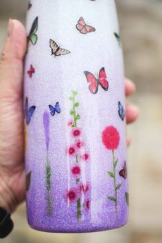 a hand holding a purple cup with flowers and butterflies painted on the outside of it