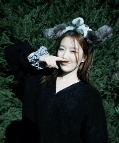 a young woman poses in front of a bush with her finger on her nose and wearing a black sweater