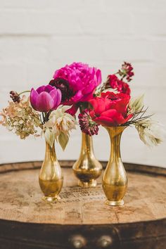 three gold vases with flowers in them sitting on a wooden table next to a brick wall