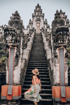 a woman in a dress and hat walking up some stairs to an old building with statues on it