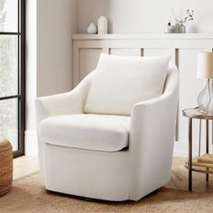 a white chair sitting in front of a window next to a table with a lamp on it