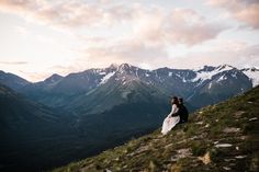 a bride and groom sitting on top of a mountain looking at the mountains in the distance