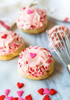 some cookies with pink frosting and sprinkles on a table next to a whisk