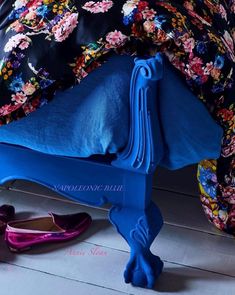 an upholstered blue bed with floral sheets and pillows on it, next to colorful slippers