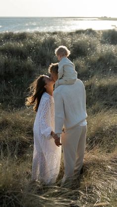 a man and woman holding a baby in their arms while walking through tall grass near the ocean