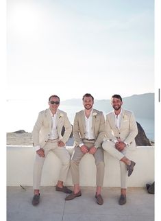 three men in suits are sitting on a white wall and smiling at the camera while wearing sunglasses