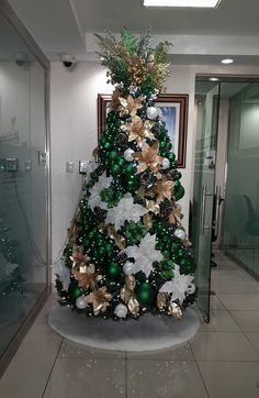 a christmas tree decorated with green and gold ornaments in an office building lobby, surrounded by glass doors