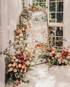 flowers and greenery are arranged in front of a mirror on a table next to a window