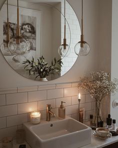 a bathroom sink sitting under a round mirror next to a vase with flowers and candles