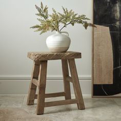 a white vase sitting on top of a wooden stool next to a painting and plant