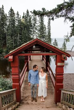 a man and woman standing on a wooden bridge near the water with pine trees in the background