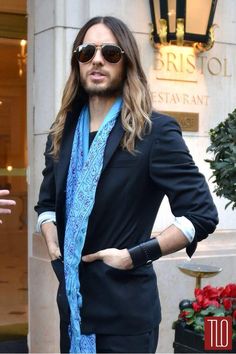 a man with long hair wearing sunglasses and a blue scarf standing in front of a building