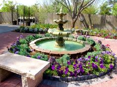 a fountain surrounded by colorful flowers in a small garden with brick pavers and stone bench