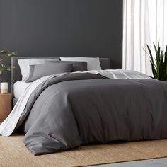 a bed with grey sheets and pillows in a room next to a plant on the floor