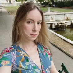 a woman sitting on a bench in front of a pond and looking at the camera