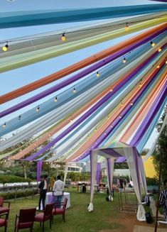 an outdoor event with colorful ribbons hanging from the ceiling and people standing under chairs on the grass