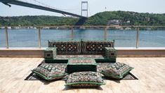 a set of couches and pillows sitting on a wooden deck overlooking the water with a bridge in the background