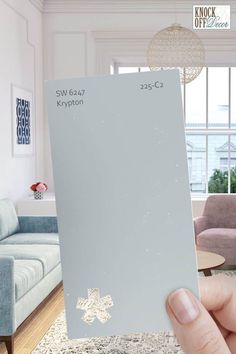 a person holding up a gray paint swatch in their living room, with the top half painted white