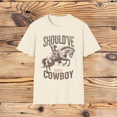 Embrace the cowboy spirit with this Should've Been a Cowboy T-Shirt! Featuring a bold, vintage-inspired design of a cowboy riding a bucking bronco, this tee captures the rugged essence of the wild west. The distressed lettering and classic western imagery make it a standout piece for any country lover's wardrobe. Key Features: Rugged Design: Detailed graphic of a cowboy on a bucking horse with the phrase "Should've Been a Cowboy" in a weathered, vintage font. Comfortable Fit: Made from soft, bre Western Style T-shirt For Rodeo, Western Style Cotton T-shirt For Rodeo, Western Style Crew Neck T-shirt For Themed Events, Western Style Crew Neck T-shirt With Graphic Print, Graphic Print Crew Neck T-shirt For Country Events, Short Sleeve T-shirt With Letter Print For Western-themed Events, Graphic Print Short Sleeve T-shirt For Country Events, Western Style Screen Print Tops For Western-themed Events, Western Tops With Screen Print For Western-themed Events