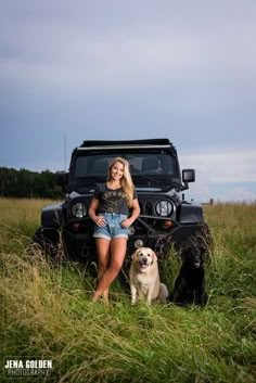 a woman standing next to two dogs in front of a black jeep on a grassy field