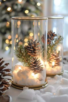 two glass jars filled with snow and pine cones on top of a table next to a christmas tree