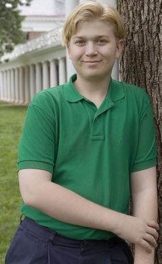 a young man leaning against a tree in front of a white house with columns on either side