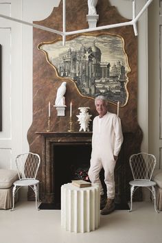 an older man standing in front of a fireplace with white chairs and a painting on the wall