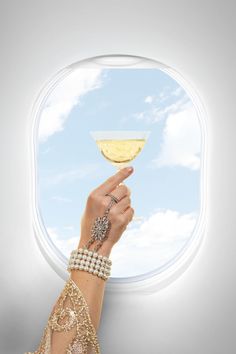 a hand holding a wine glass in front of an airplane window with the sky and clouds behind it