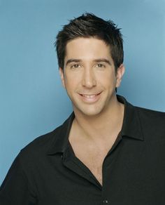 a man in black shirt smiling at the camera with blue background and white wall behind him