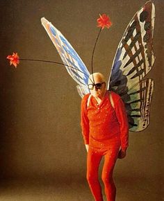 a man dressed as a butterfly with sunglasses on his head and red pants, holding a flower in one hand