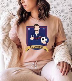 a woman sitting down wearing a t - shirt with the image of a man holding a soccer ball