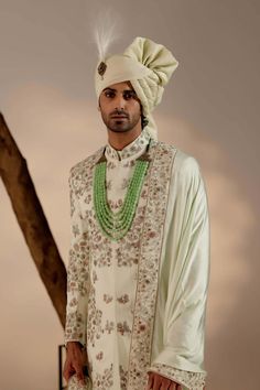 a man wearing a white outfit and green beaded necklaces standing in front of a tree