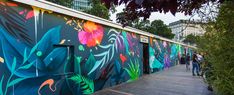 people walking past a colorfully painted wall on a boardwalk in the city, next to trees