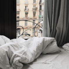 an unmade bed sitting in front of a window next to a tall building with lots of windows