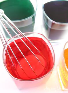 three mixing bowls with different colored liquids in them and one whisk on top